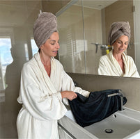 Woman in a bathrobe with a towel on her head, standing by a sink in a bathroom.